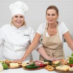 Two female chefs with variety of fresh ingredients in a kitchen setting, showcasing culinary preparation.